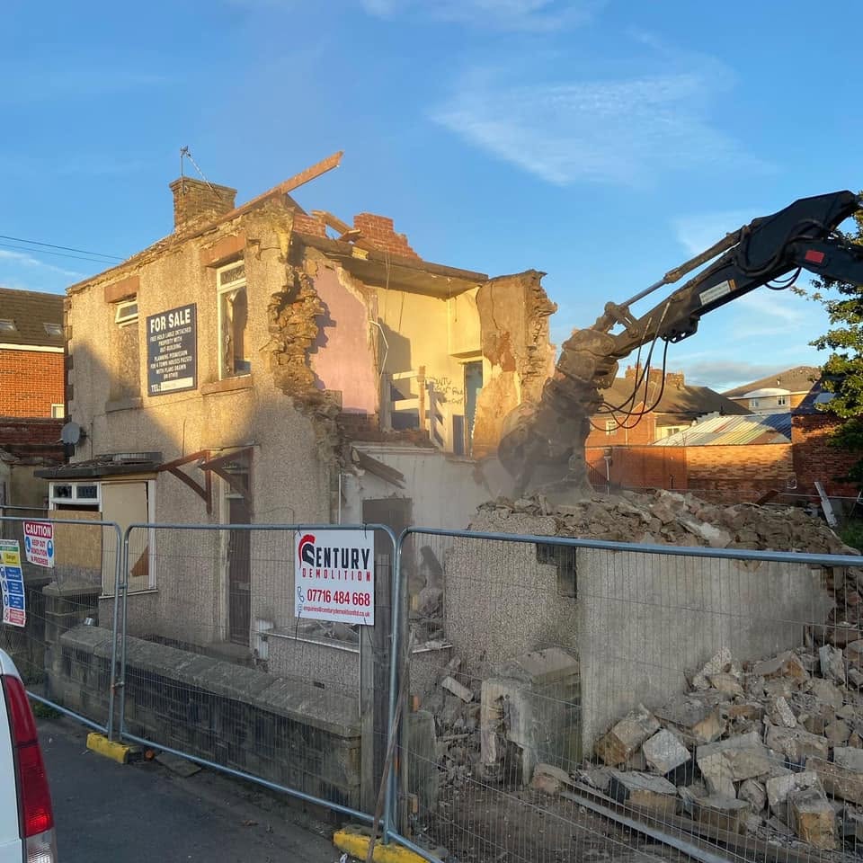 Century Demolition excavator taking down a semi-detached house behind site fencing displaying the company board